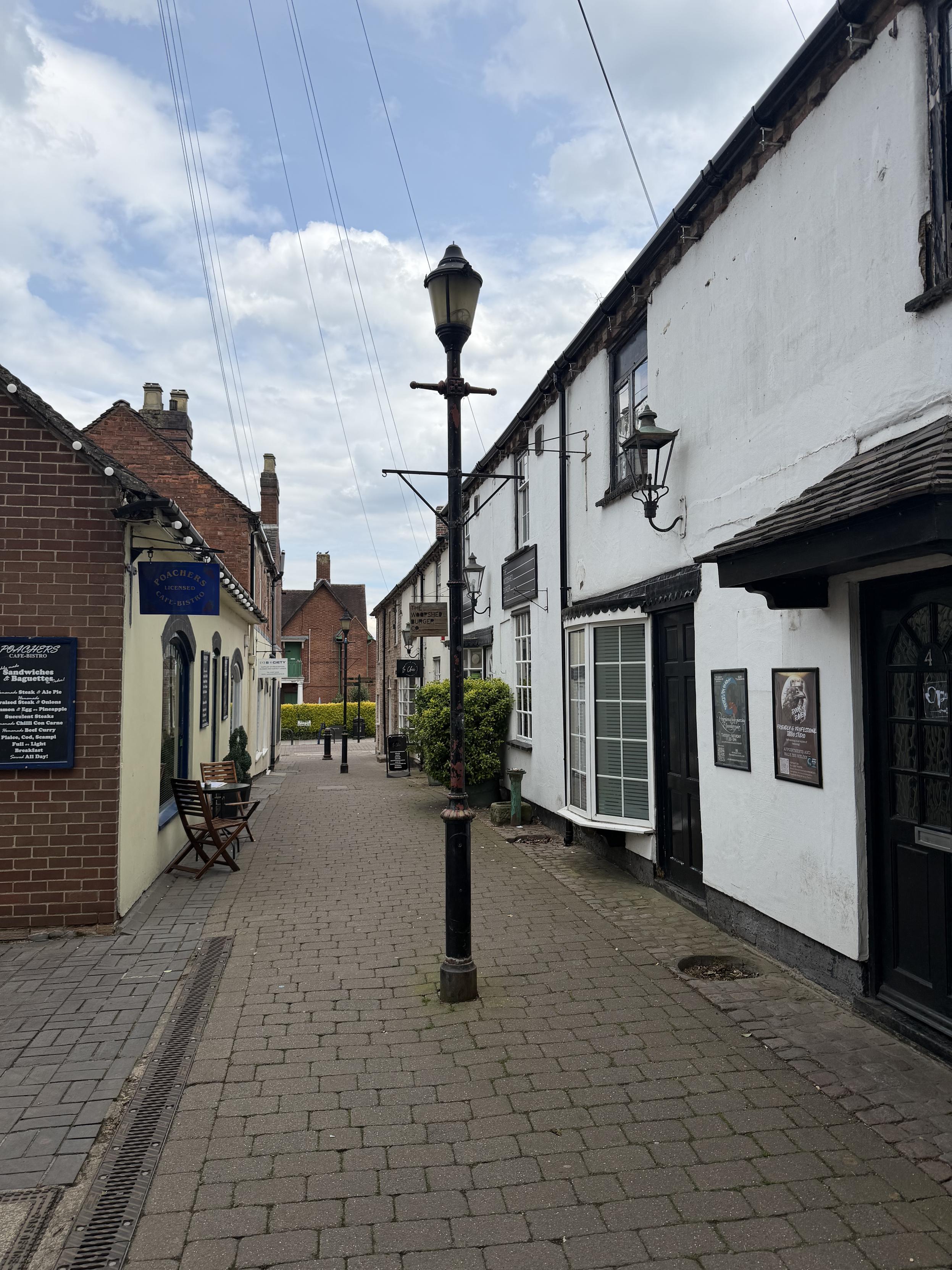 A quiet alleyway with cobblestone pavement, lined with small buildings with white and brick facades, and a streetlamp in the center.