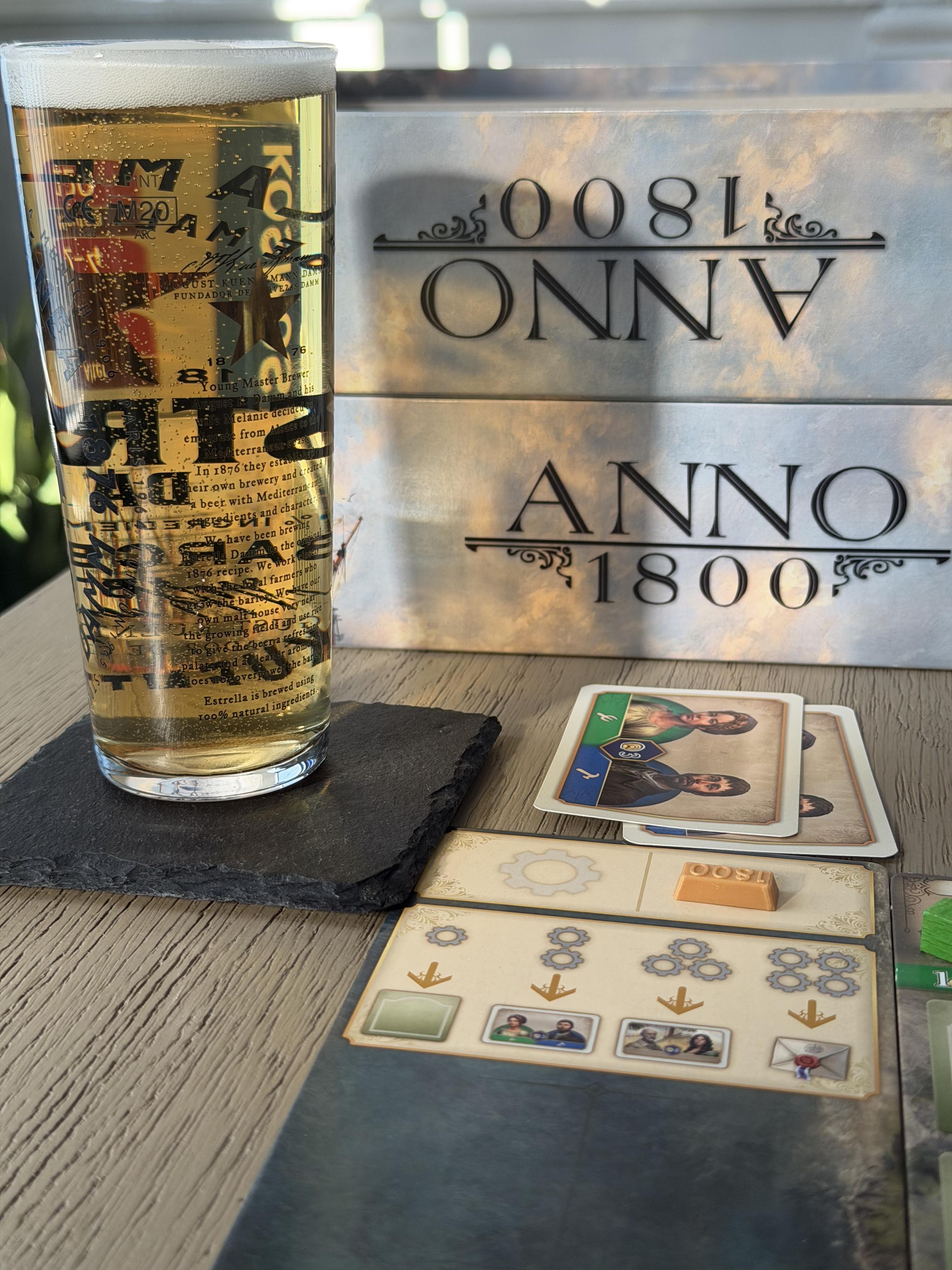 A glass of beer on a slate coaster, with Anno 1800 board game pieces and cards on a wooden table, and the Anno 1800 box in the background.