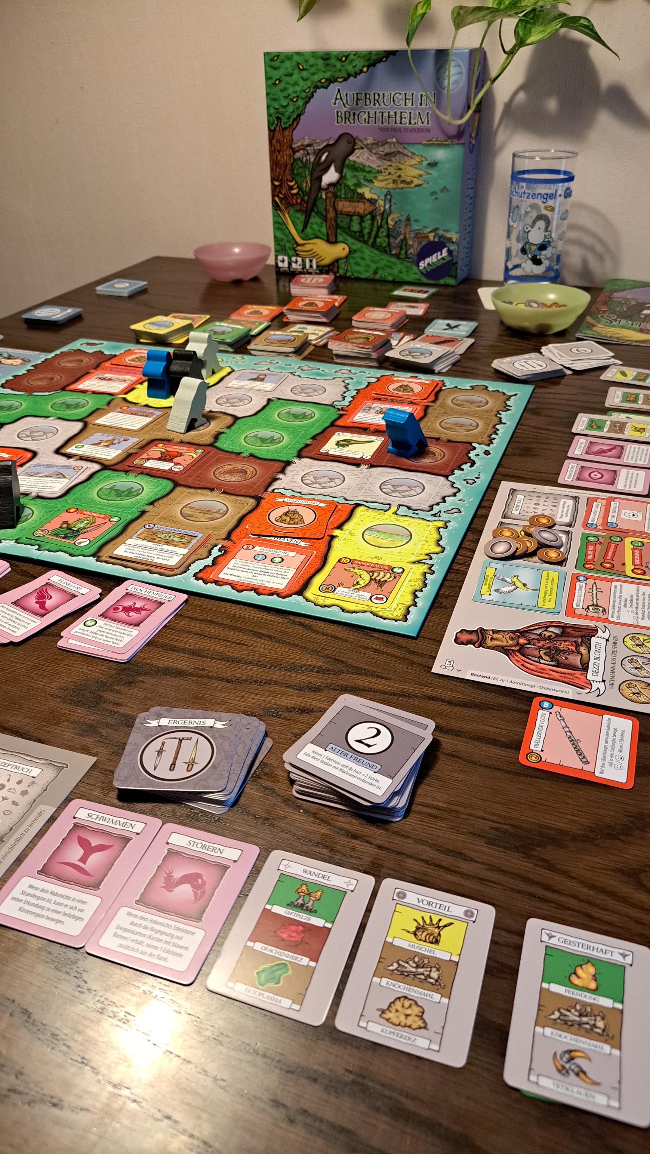 The game on a wooden table: a main board, player boards, lots of cards, player meeples, and tokens. In the background, there is the game box lid showing 2 birds sitting on a tree branch and looking at landscape.